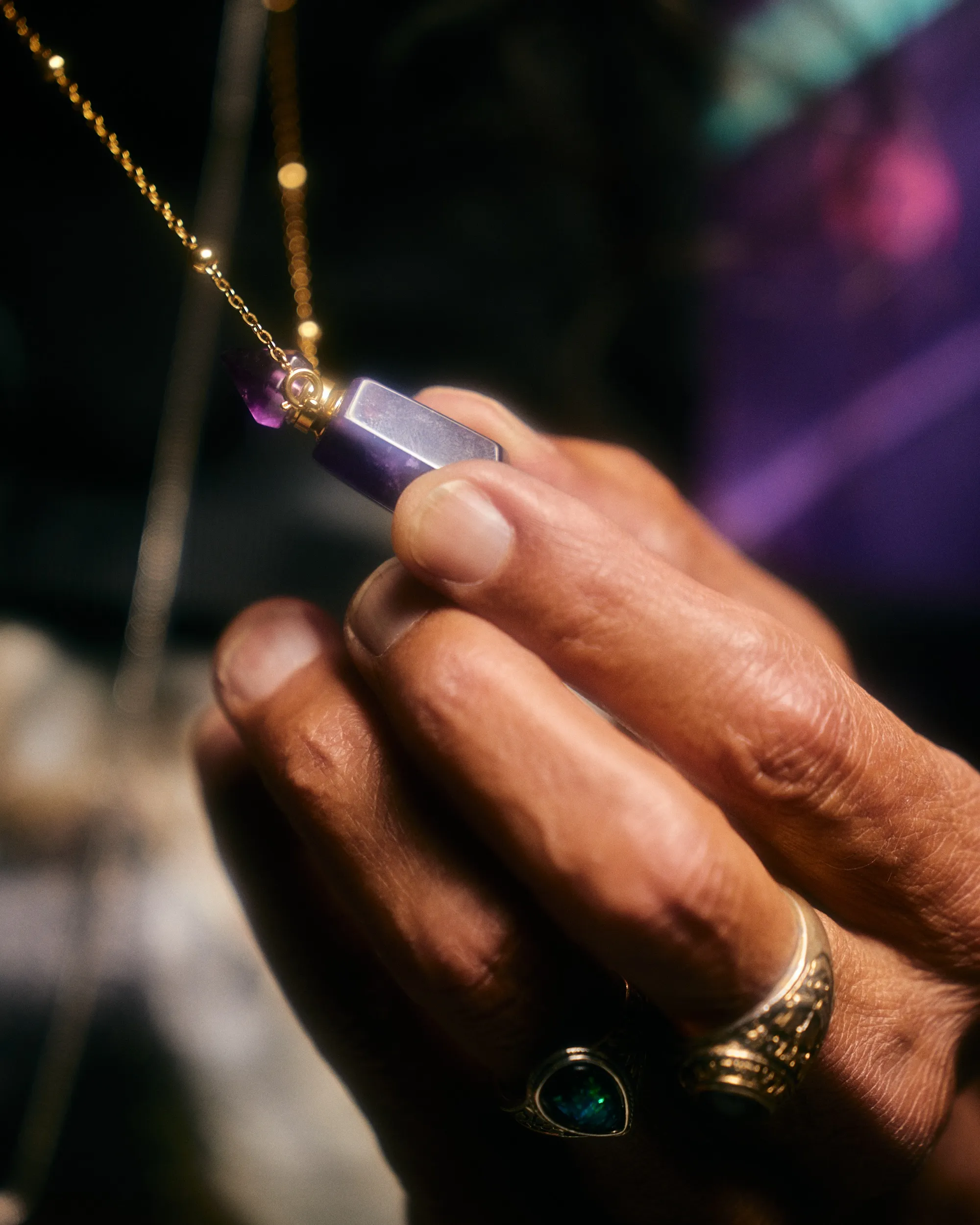 Close-up of fingers with rings holding a purple crystal pendant on a gold chain