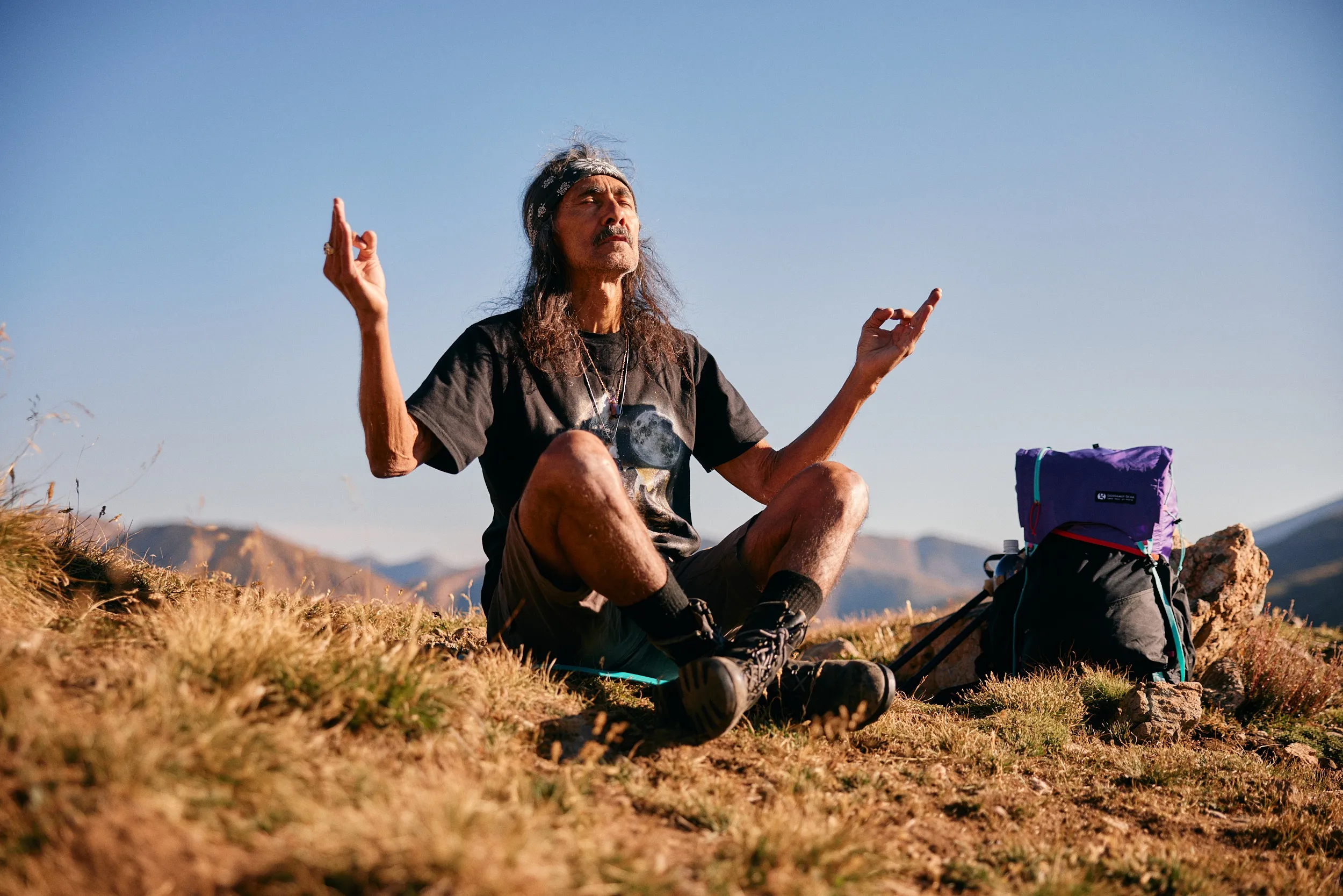 Hiker meditating cross-legged on a grassy alpine ridge beside a purple pack