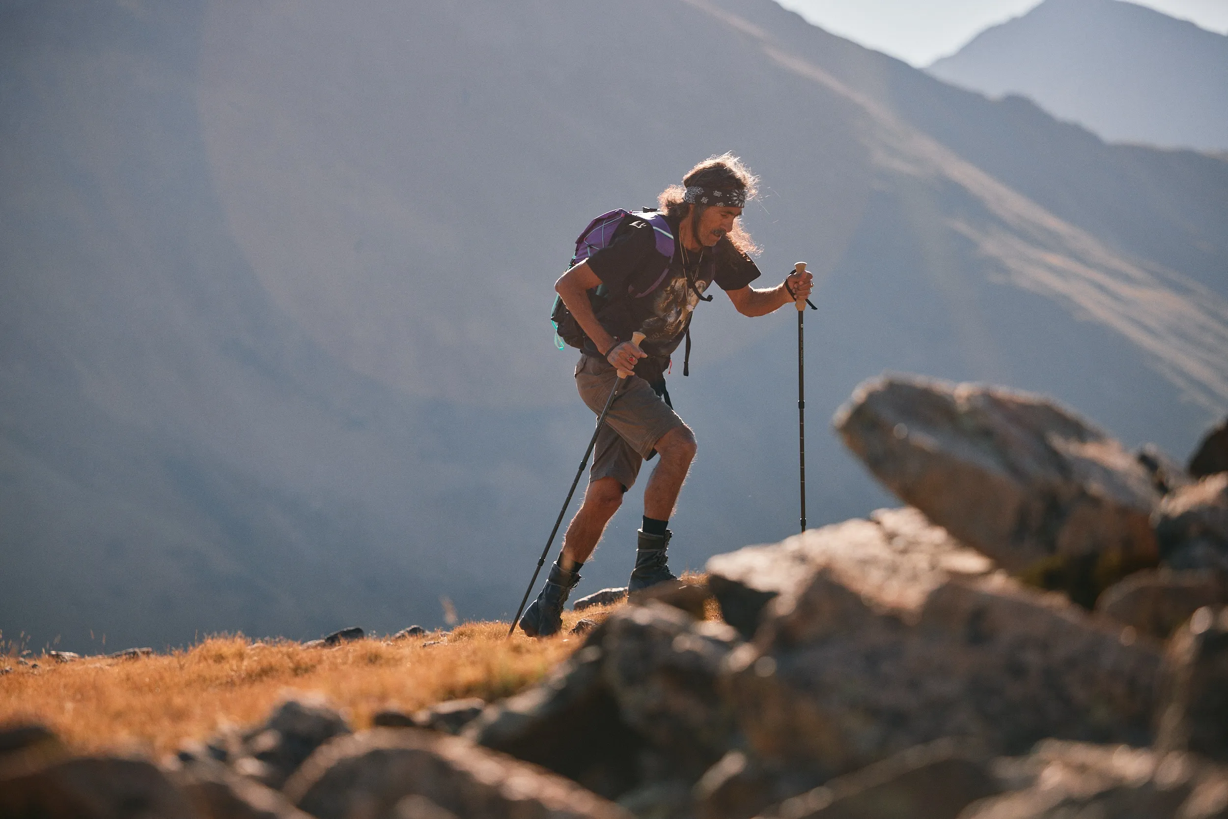 Hiker with a purple pack and trekking poles climbing over rocks on a sunny mountainside